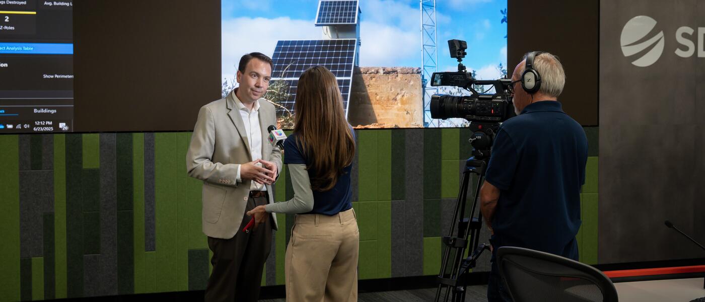 A media interview takes place at SDG&E's WCRC with illuminated screens in the background.