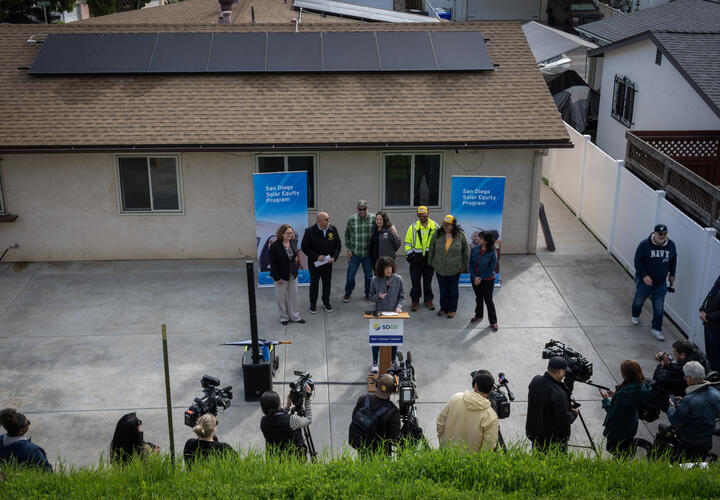 A group of people participate in a press event, in the backyard of a residential home.