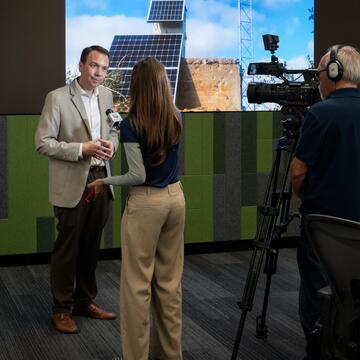 A media interview takes place at SDG&E's WCRC with illuminated screens in the background.