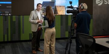 A media interview takes place at SDG&E's WCRC with illuminated screens in the background.