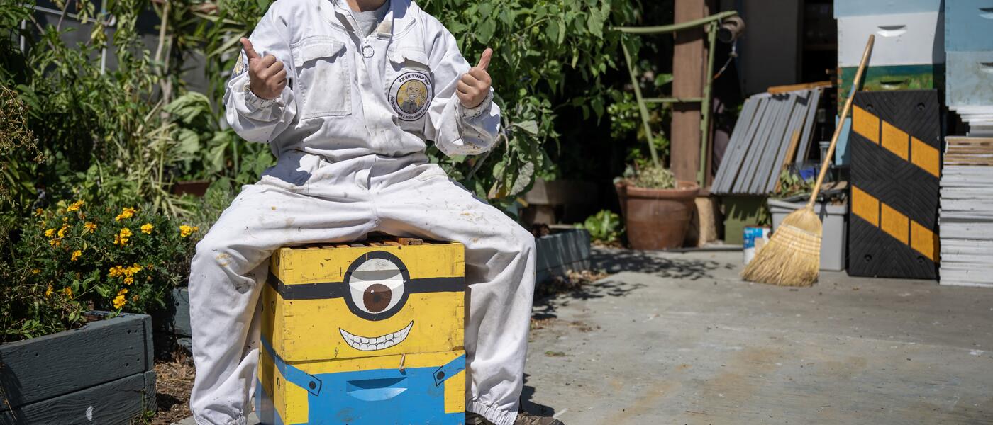 Dan the Bee Man poses for a photo, sitting on a colorful wood box and giving a thumbs up sign. 
