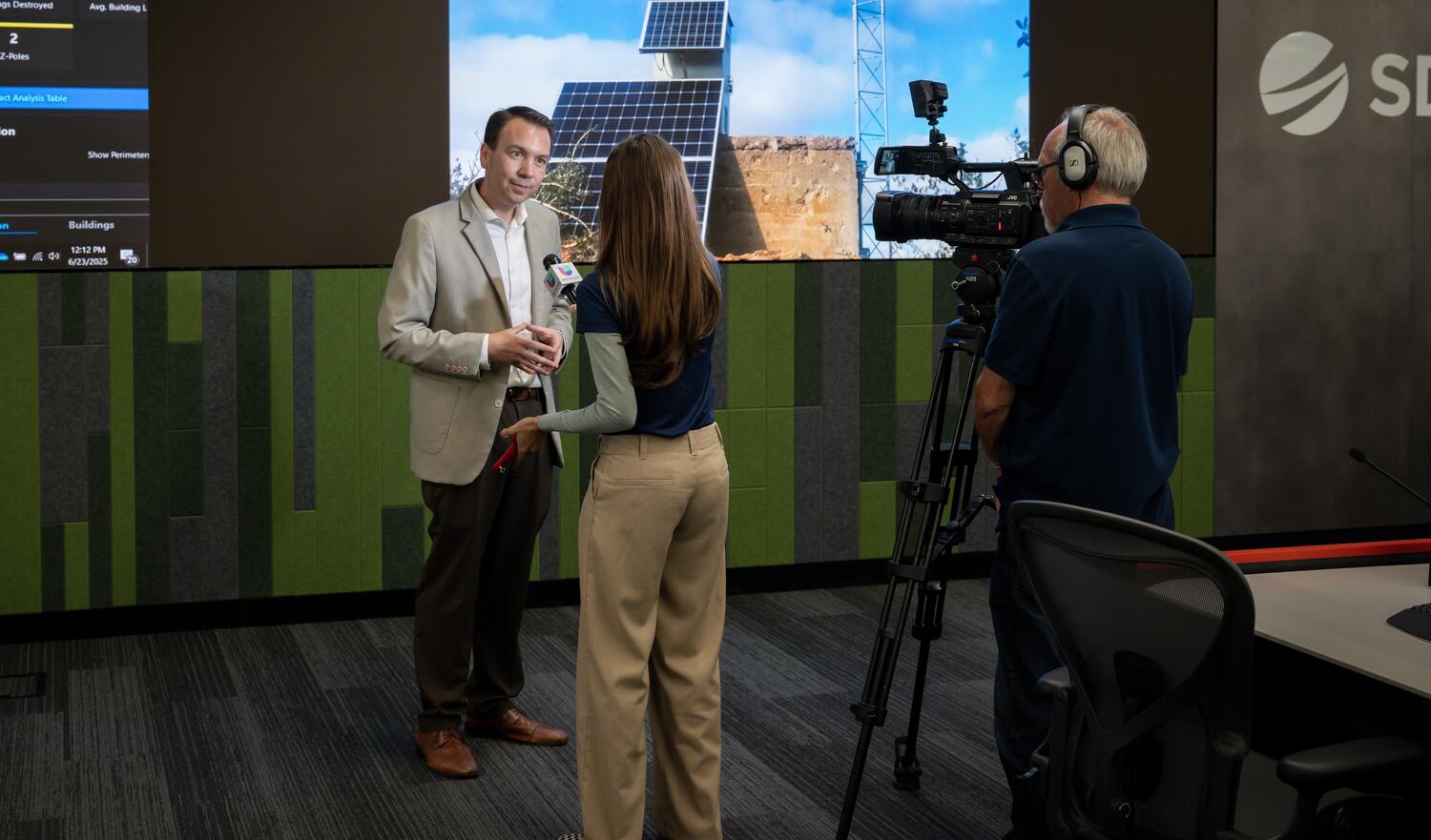 A media interview takes place at SDG&E's WCRC with illuminated screens in the background.