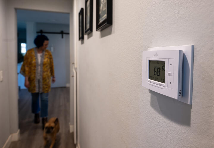 A white, digital thermostat on the wall, with a woman and her dog walking down the hallway in the background