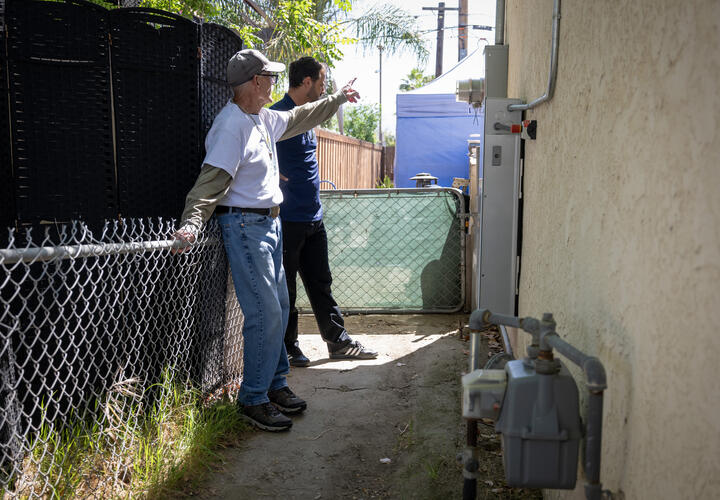 two men stand on the side of a home, near a electric meter box and battery storage