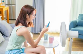 A woman sitting on a couch, looking at her mobile phone in front of a fan